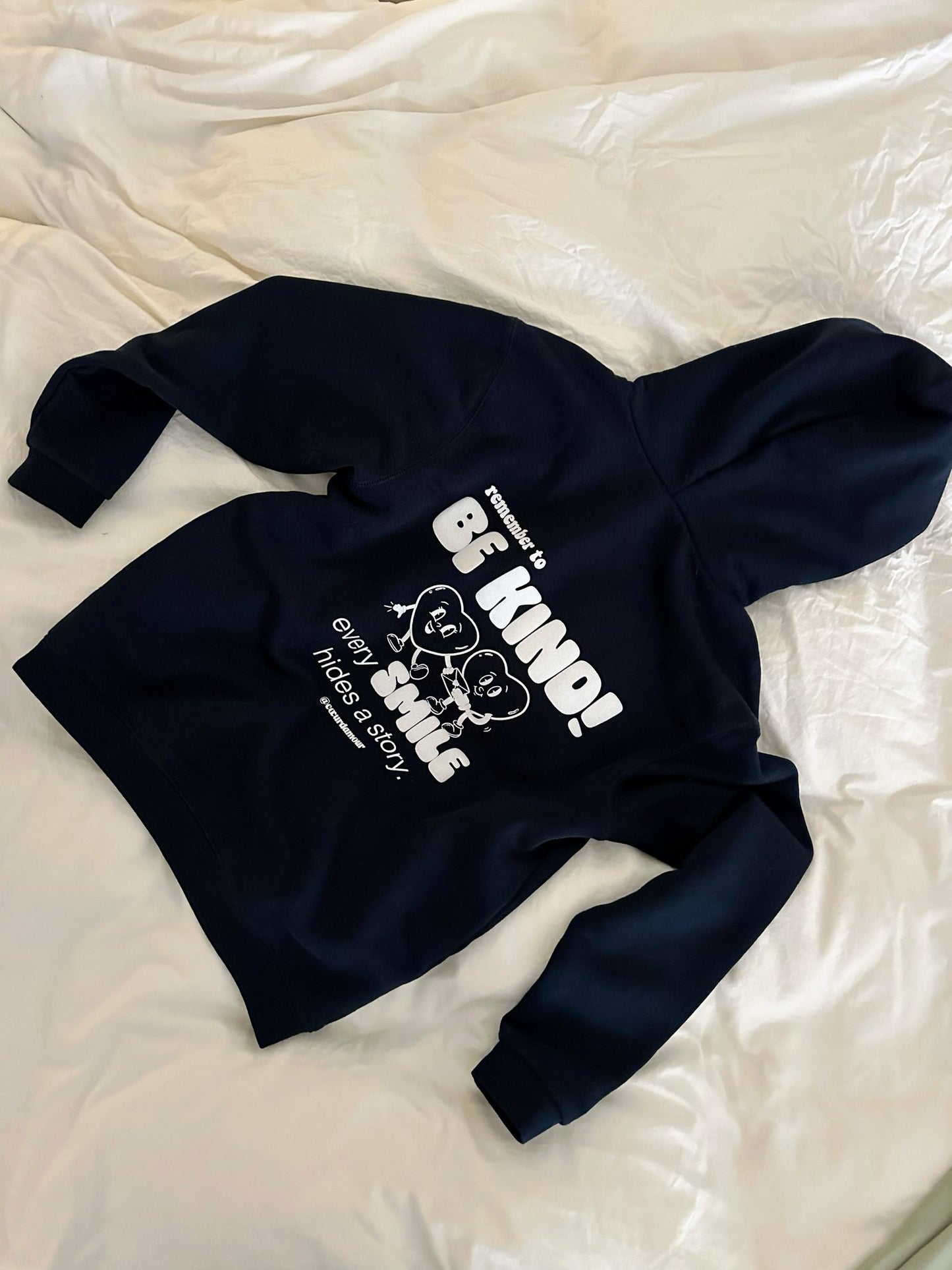 Hoodie liegt auf einem weißen Laken auf dem Boden, weißer Backprint "remember to BE KIND! every SMILE hides a story"