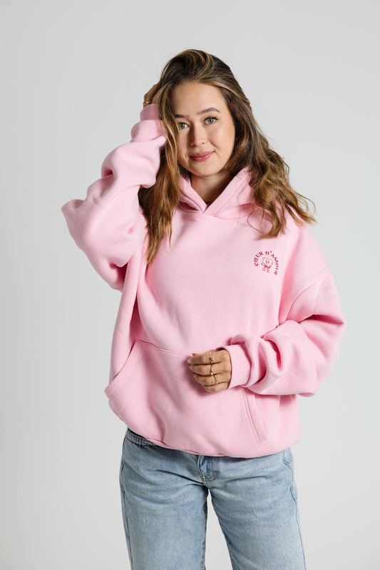 Frau mit offenen Haaren trägt ein rosa Oversize Hoodie mit rotem "Coeur d'Amour" Aufdruck auf der linken Brust. Dazu eine hellblaue Jeans vor neutralem Hintergrund.