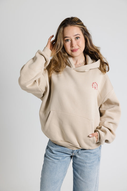 Beiger Oversize-Hoodie mit rotem Aufdruck " Coeur d'amour auf der linken Brust, getragen von einer Frau mit offenen Haaren.