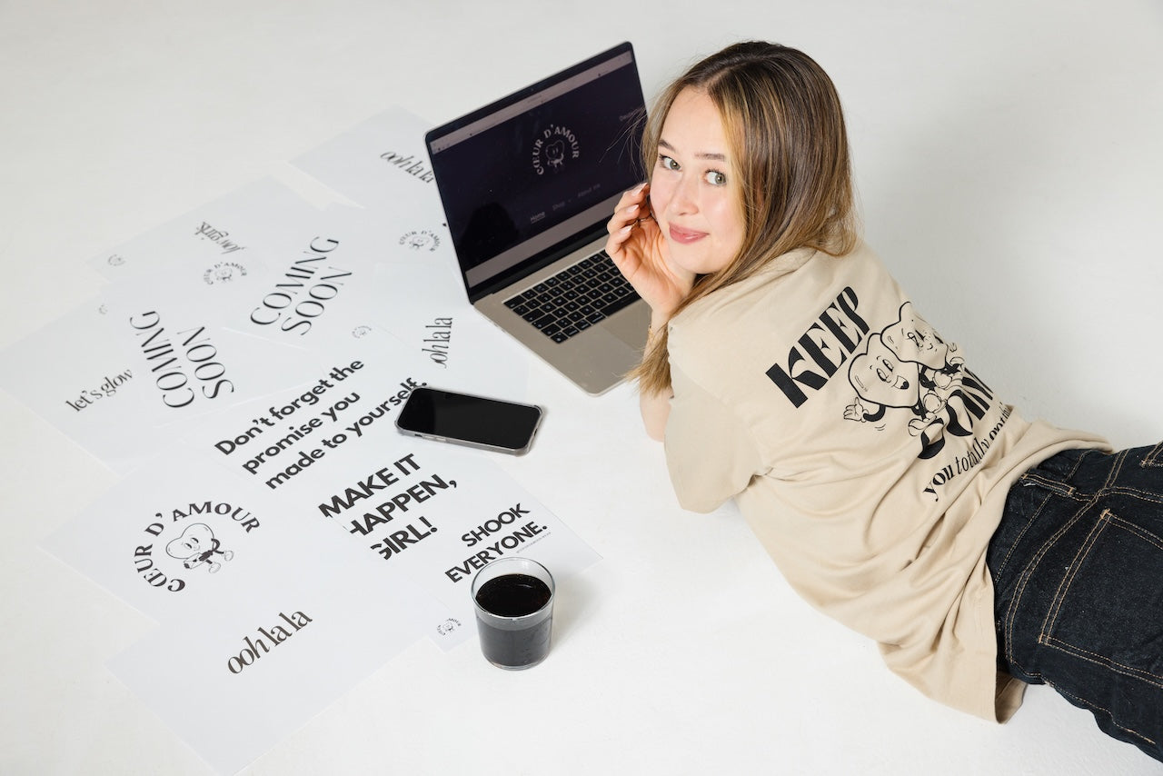 Junge Frau liegt lächelnd auf dem Boden vor einem geöffneten Laptop mit der Website „COEUR D’AMOUR“. Sie trägt ein beigefarbenes T-Shirt mit großem Rückendruck: Comicfiguren und der Schriftzug „KEEP GOING – you totally got this“. Um sie herum liegen mehrere Blätter mit typografischen Botschaften wie „COMING SOON“, „MAKE IT HAPPEN, GIRL!“ oder „Don’t forget the promise you made to yourself“. Ein Smartphone und ein Becher mit dunklem Getränk liegen ebenfalls daneben. Heller, minimalistischer Hintergrund.