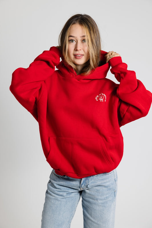 Frau von vorne, trägt ein roten Oversize Hoodie mit weißem "Coeur d'Amour" Logo auf der linken Brust. Dazu eine hellblaue Jeans vor neutralem Hintergrund.