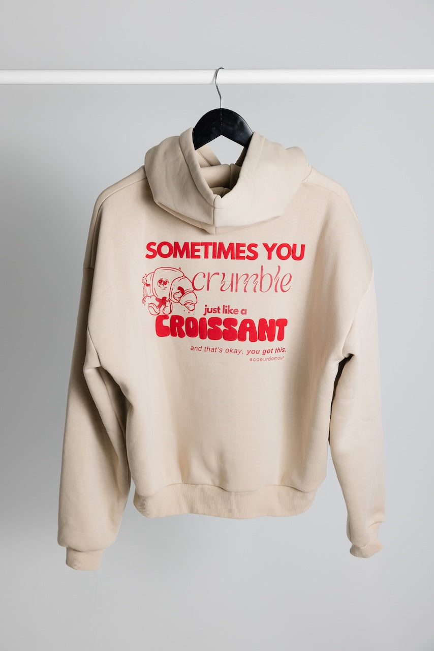 Beiger Oversize-Hoodie mit rotem Aufdruck "Sometimes you crumble just like a croissant" und Croissant-Grafik, hängend an einem Kleiderbügel vor neutralem Hintergrund.