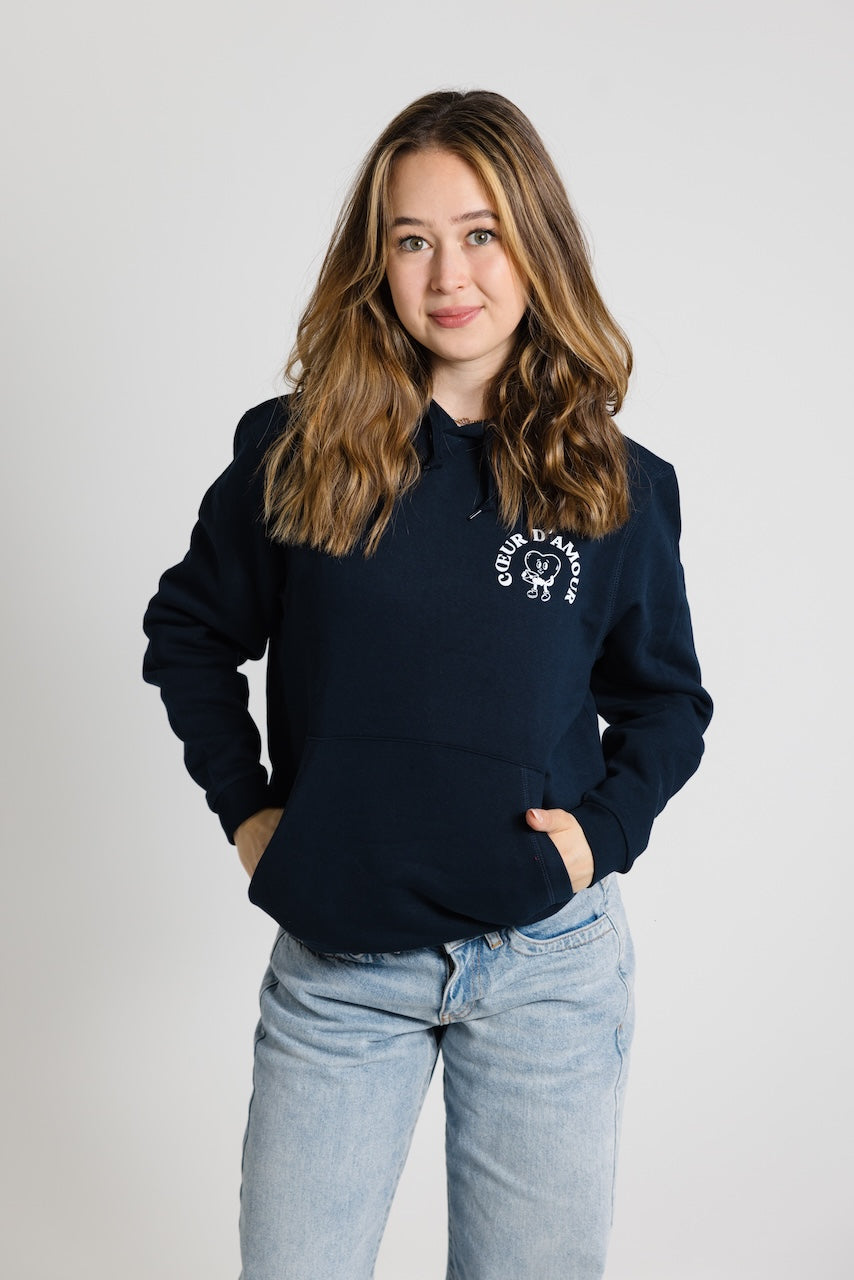 Frau von vorne, trägt ein dunkelblauen Oversize Hoodie mit weißem "Coeur d'Amour" Logo auf der linken Brust. Dazu eine hellblaue Jeans vor neutralem Hintergrund.