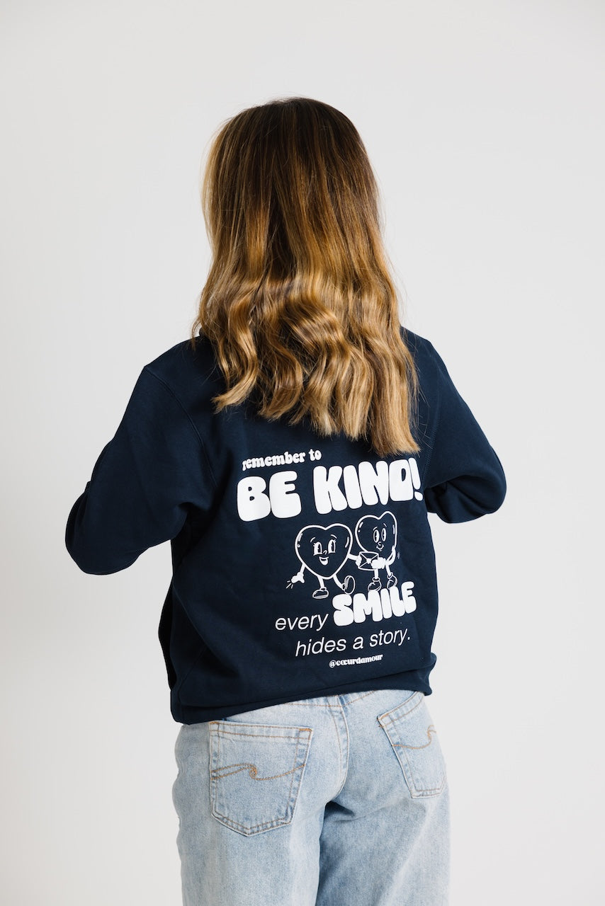 Frau von hinten, trägt ein dunkelblauen Oversize Hoodie mit weißem Backprint "remember to BE KIND every SMILES hides a Story". Dazu eine hellblaue Jeans vor neutralem Hintergrund.