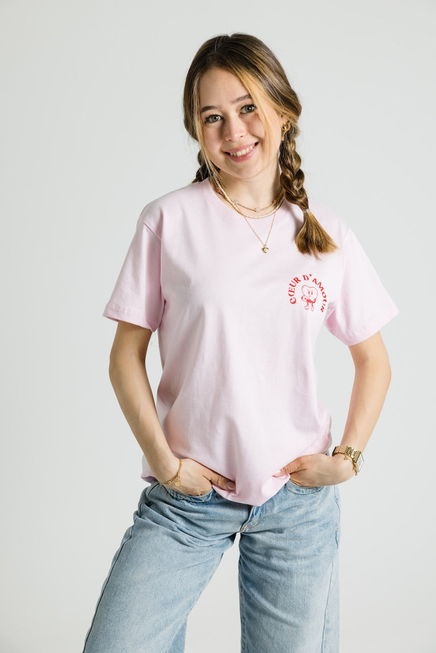 Eine Person trägt ein rosafarbenes T-Shirt mit kleinem Brustprint: ein Herzcharakter mit dem Schriftzug „COEUR D’AMOUR“ in Rot. Das T-Shirt hat eine lockere Passform.