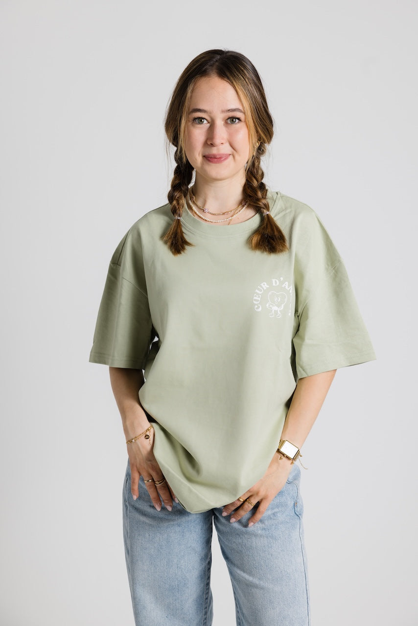 Frau mit geflochtenen Zöpfen trägt ein hellgrünes Oversize-T-Shirt mit weißem Aufdruck ‚Cœur d’Amour‘ und blauer Jeans vor neutralem Hintergrund.