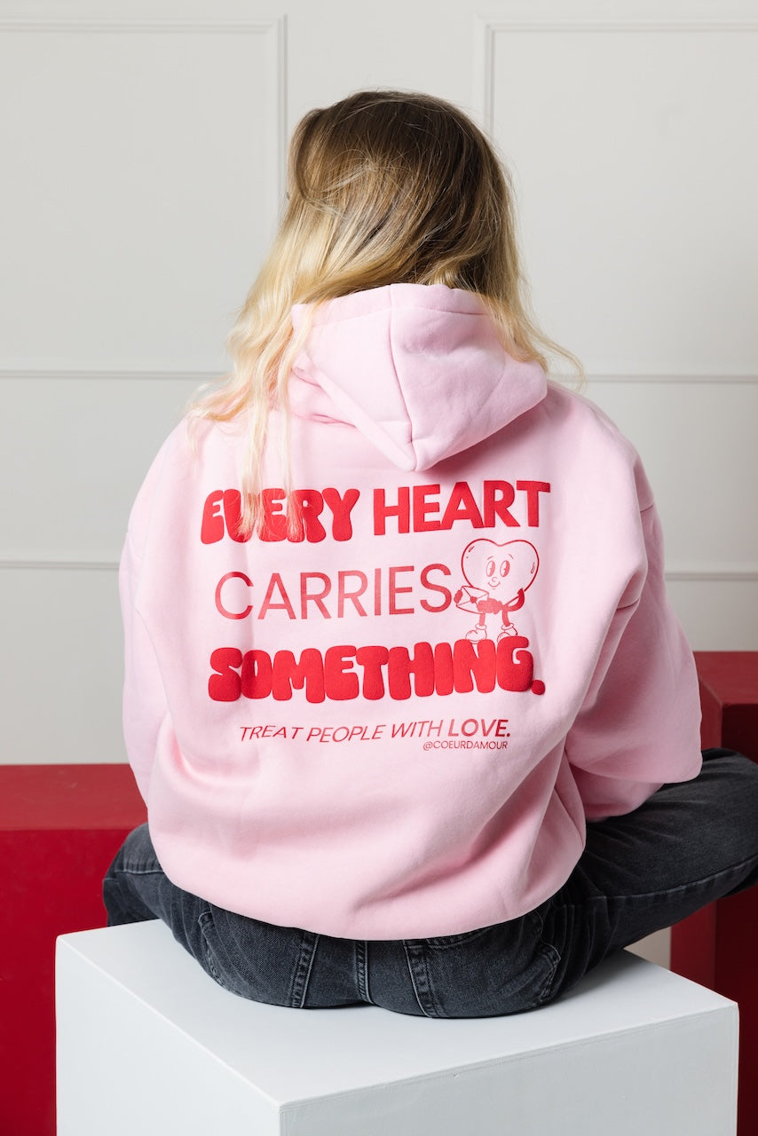 Frau sitzend von hinten, trägt einen rosa Oversize Hoodie mit rotem "Every Heart carries something that people with love" Aufdruck
