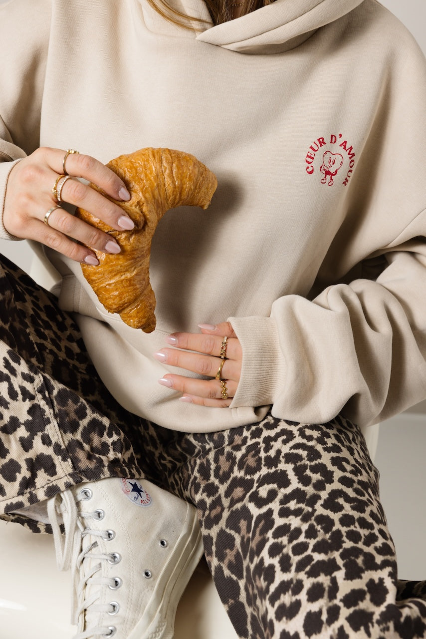 Frau sitzend mit einem Croissant, Close-up, trägt einen beigefarbenen Hoodie mit rotem "Coeur d'amour Aufdruck auf der linken Brust.