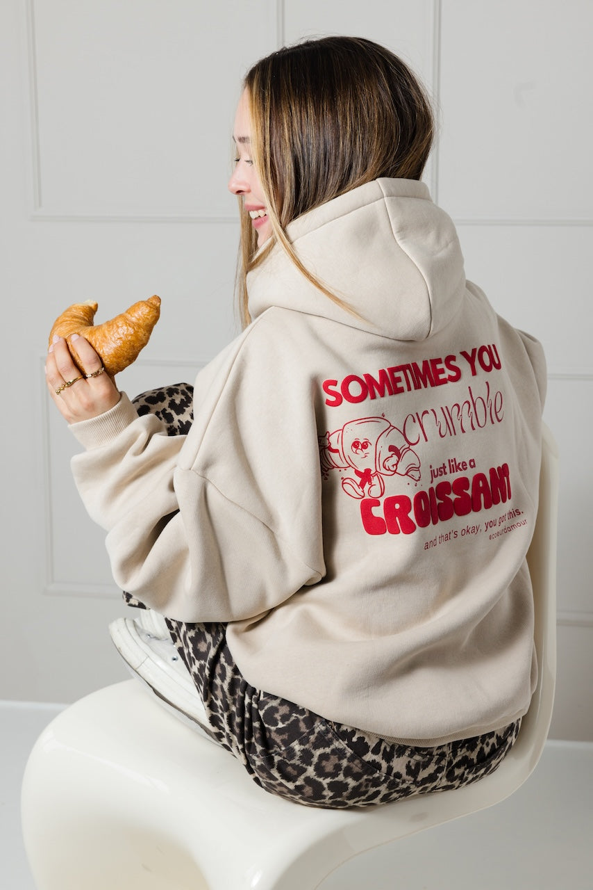 Frau sitzend von hinten trägt einen beigefarbenen Hoodie mit rotem Croissant-Motiv und Spruch ‚Sometimes you crumble just like a croissant‘, mit einem Croissant in der linken Hand haltend