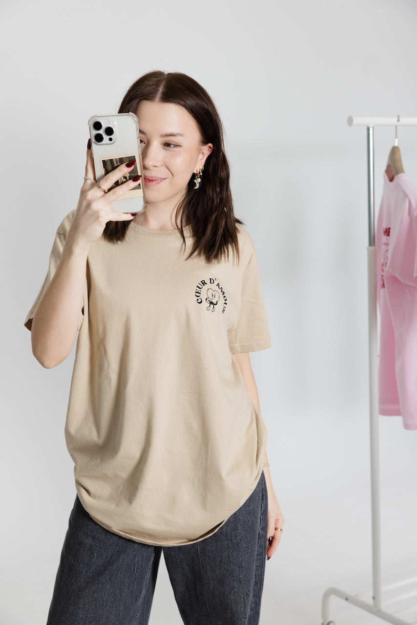 Frau mit beigem T-Shirt mit schwarzen "Coeur d'Amour" macht ein Spiegelfoto, trägt dunkle Hose und steht vor Kleiderständer mit rosa Shirt im Hintergrund.