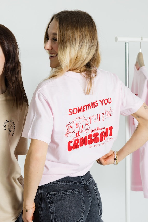 Zwei Personen von hinten im Gespräch. Eine trägt das rosafarbene T-Shirt mit dem roten Croissant-Motiv, die andere ein beiges Shirt mit dem „COEUR D’AMOUR“-Brustprint in Schwarz.