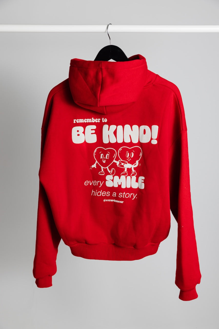 Oversize Hoodie von hinten hängt an einem Kleiderbügel vor neutralem Hintergrund, darauf ein weißer Backprint "remember to BE KIND every SMILES hides a Story".