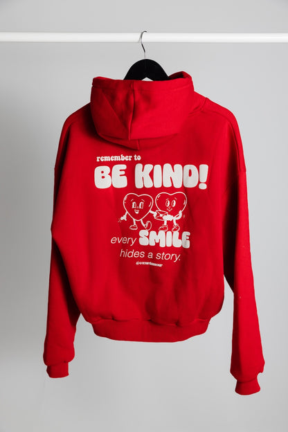 Oversize Hoodie von hinten hängt an einem Kleiderbügel vor neutralem Hintergrund, darauf ein weißer Backprint "remember to BE KIND every SMILES hides a Story".