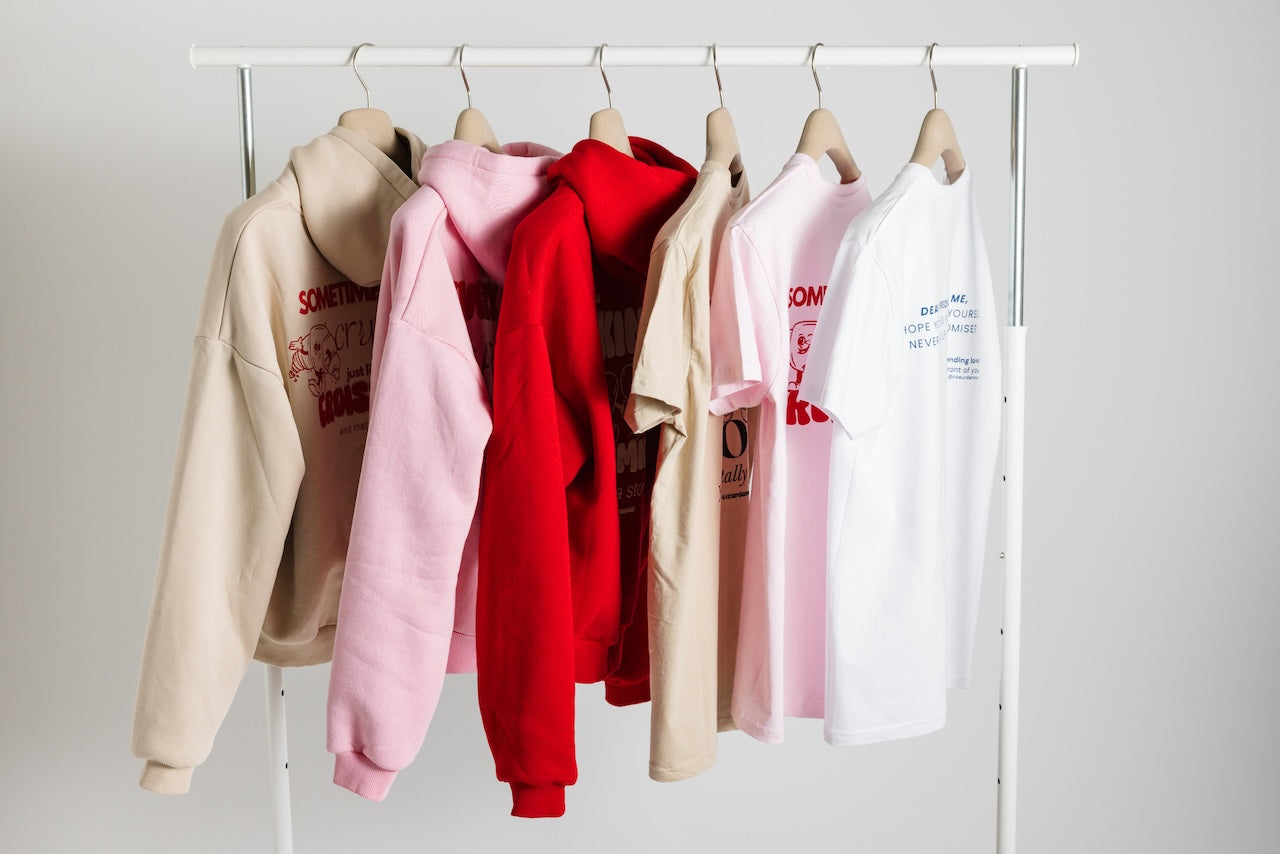 Kleiderstange mit sechs Kleidungsstücken in hellen Farben, von links nach rechts: beigefarbener Hoodie mit rotem Rückendruck, rosa Hoodie mit rotem Aufdruck, roter Hoodie, beigefarbenes T-Shirt mit schwarzem Druck, rosa T-Shirt mit rotem Motiv und weißes T-Shirt mit blauem Druck. Alle Teile hängen ordentlich auf Holzkleiderbügeln vor neutralem, hellem Hintergrund.
