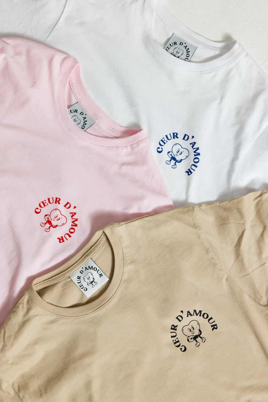 Drei T-Shirts flach nebeneinander liegend, jeweils mit kleinem Frontprint links auf der Brust. Das weiße Shirt zeigt einen blauen Aufdruck eines Comic-Herz-Charakters mit dem Schriftzug „COEUR D’AMOUR“ im Halbkreis. Das rosa Shirt trägt den gleichen Print in Rot, das beige Shirt in Schwarz. Alle Shirts haben einen gerippten Rundhalsausschnitt und ein Nackenlabel mit dem gleichen Designmotiv.