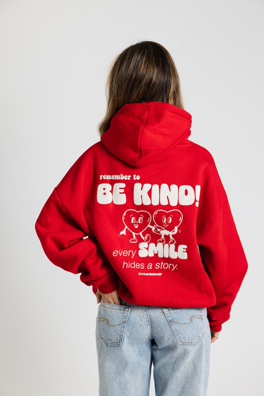 Frau von hinten, trägt ein roten Oversize Hoodie mit weißem Backprint "remember to BE KIND every SMILES hides a Story". Dazu eine hellblaue Jeans vor neutralem Hintergrund.