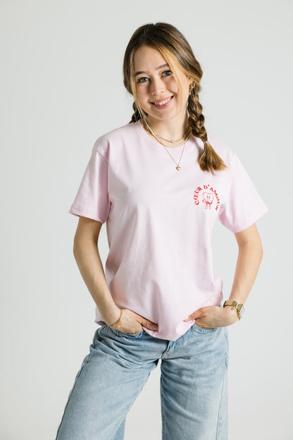 Eine Person trägt ein rosafarbenes T-Shirt mit kleinem Brustprint: ein Herzcharakter mit dem Schriftzug „COEUR D’AMOUR“ in Rot. Das T-Shirt hat eine lockere Passform.