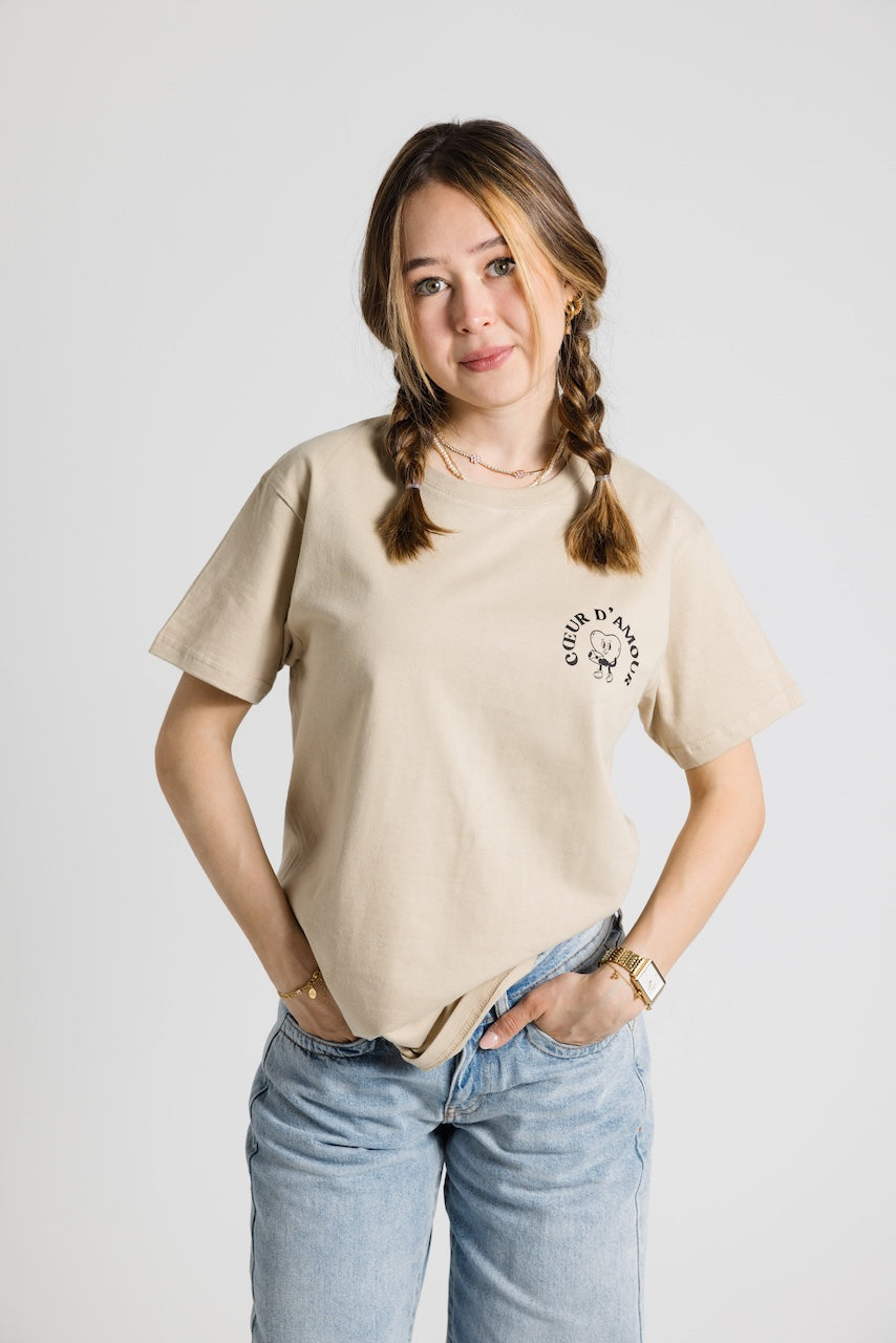 Frau mit Zöpfen trägt ein beiges Oversize T-Shirt mit schwarzem "Coeur d'Amour" Logo auf der linken Brust. Dazu eine hellblaue Jeans vor neutralem Hintergrund
