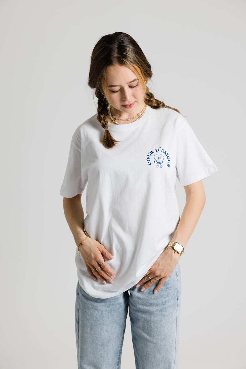 Frau von vorne trägt ein weißes Shirt mit blauem "Coeur d'amour" Aufdruck auf der linken Brust. Dazu eine hellblaue Jeans