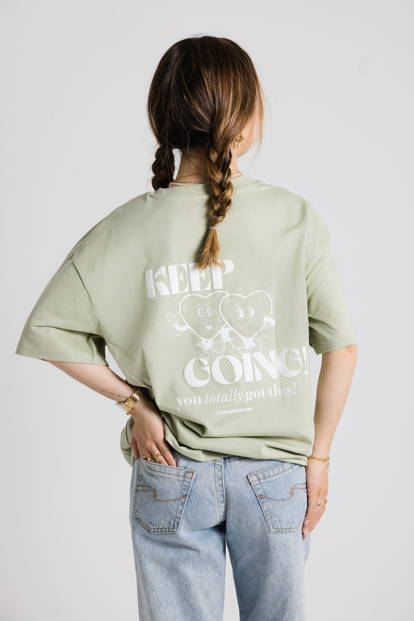 Frau mit geflochtenen Zöpfen von hinten, trägt ein hellgrünes Oversize-Shirt mit weißem Aufdruck ‚Cœur d’Amour‘ und blauer Jeans vor neutralem Hintergrund.