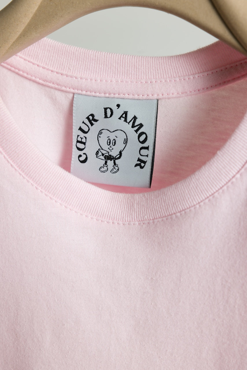  Nahaufnahme des Halsausschnitts eines rosafarbenen T-Shirts auf einem Kleiderbügel. Sichtbar ist das weiße Etikett mit schwarzem Logo „COEUR D’AMOUR“ und Herzfigur mit Brief.