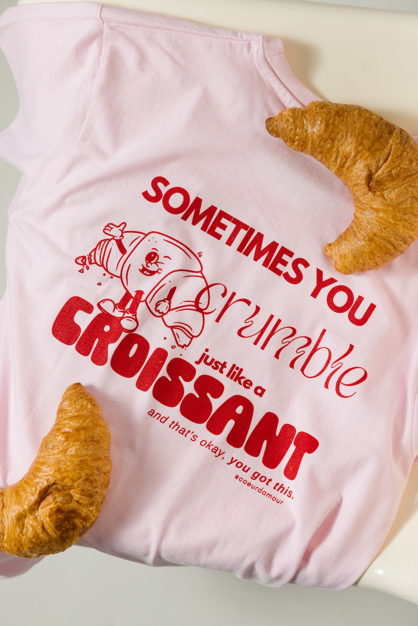 Flach liegendes rosafarbenes T-Shirt mit Croissant-Print in Rot. Aufdruck zeigt einen Comic-Croissant mit Text: „SOMETIMES YOU crumble just like a CROISSANT and that’s okay, you got this.“ Zwei echte Croissants liegen dekorativ daneben.